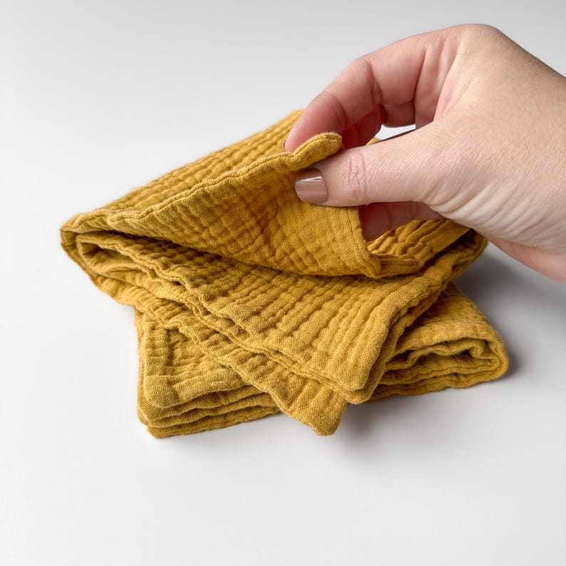 May include: A mustard yellow muslin cloth featuring a soft, multi-layered crinkled texture. The fabric is folded into several layers, showcasing the lightweight, breathable weave of the material. The square-shaped textile has finished edges.