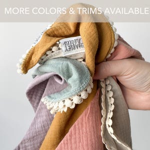May include: A collection of layered muslin cloths in mustard yellow, sage green, dusty rose, and lavender, each featuring a white pom-pom trim along the edges. A white fabric label with the black text "charley charles" is attached to the mustard cloth.