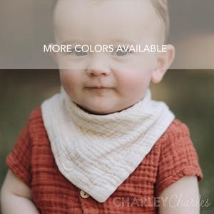 May include: A baby wearing a cream-colored bandana bib over a rust-colored short-sleeved shirt. The bib is made of a textured fabric. The text "MORE COLORS AVAILABLE" is displayed across the top of the image. The text "CHARLEY Charles" is displayed at the bottom.