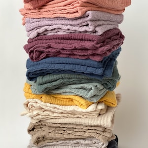 May include: A vertical stack of folded muslin cloths in a variety of colors including terracotta, dusty pink, lavender, deep plum, navy blue, sage green, mustard yellow, cream, beige, light gray, and charcoal black, all featuring a crinkled texture.