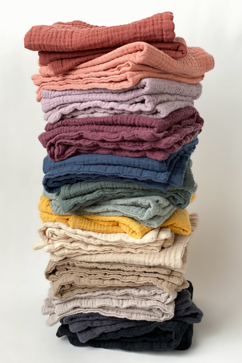 May include: A vertical stack of folded muslin cloths in a variety of colors including terracotta, dusty pink, lavender, deep plum, navy blue, sage green, mustard yellow, cream, beige, light gray, and charcoal black, all featuring a crinkled texture.