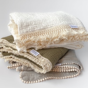May include: A stack of three folded muslin blankets in cream, olive green, and gray. The cream and olive blankets feature a cream-colored fringe trim, while the gray blanket has a cream pom-pom trim. Each blanket has a white Charley Charles label.