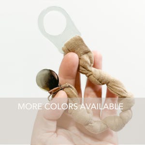 May include: A tan fabric pacifier clip featuring a translucent white plastic ring at one end and a metallic silver circular clip at the other. The fabric is gathered and twisted along the length of the strap. The text MORE COLORS AVAILABLE is visible.