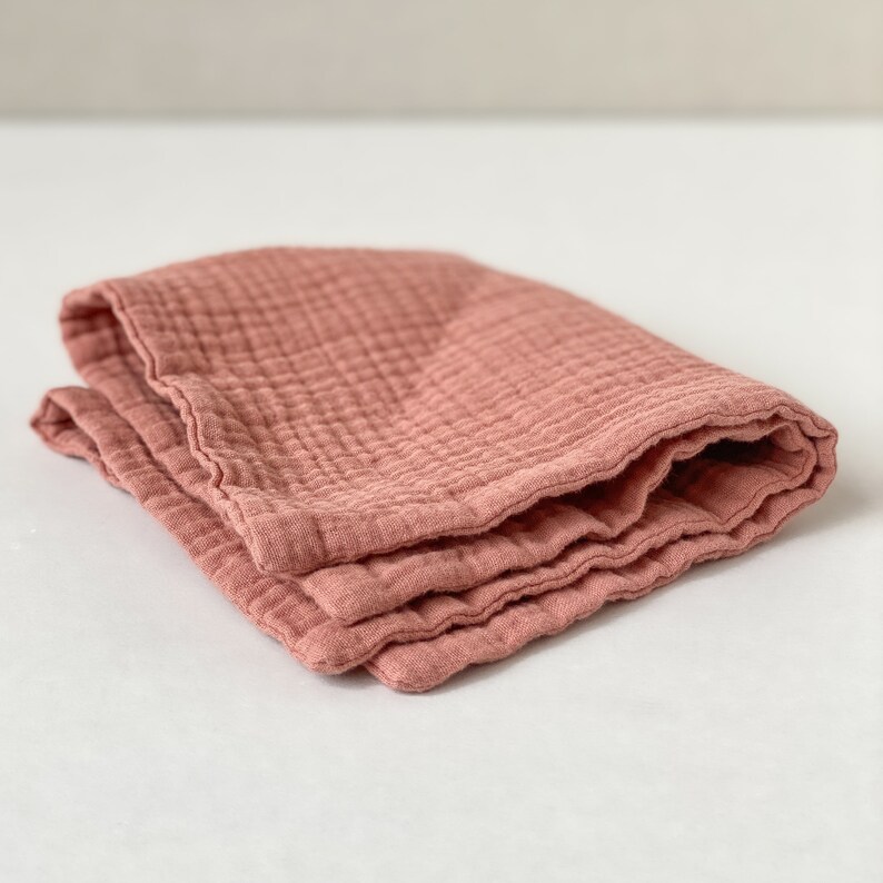 May include: A folded square of dusty rose colored muslin fabric featuring a distinct crinkled, multi-layered texture. The textile displays a grid-like pattern formed by the stitching of the layers, creating a soft, puckered appearance throughout the material.