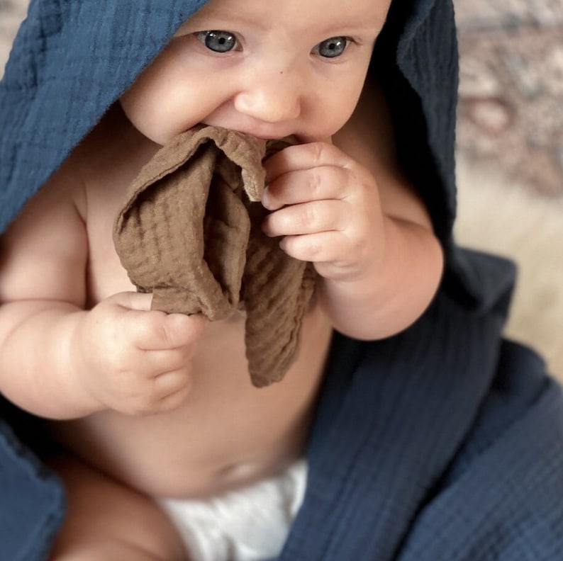 May include: A dark blue hooded muslin towel draped over the head and shoulders, paired with a small, textured brown muslin cloth held by the hands. The fabric features a soft, crinkled texture characteristic of multi-layered cotton muslin material.