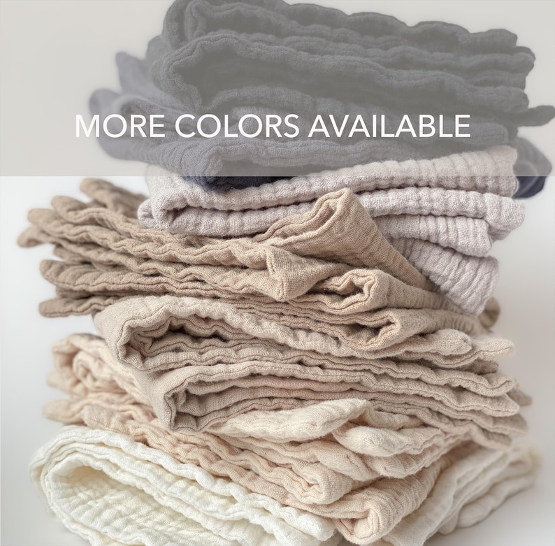 May include: A stack of folded muslin cloths in various neutral shades including white, cream, beige, tan, and dark gray. The fabric has a crinkled, textured appearance. The text MORE COLORS AVAILABLE is printed in white across the top of the stack.
