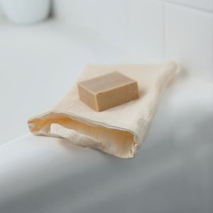 May include: A light brown bar of soap rests on a cream-colored washcloth. The washcloth is folded and placed on a white surface, likely a bathroom sink or tub. The soap is rectangular and appears to be unscented.