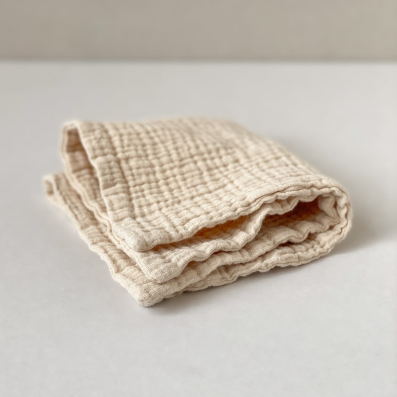 May include: A folded square cloth made of beige muslin fabric with a visible crinkled, multi-layered texture. The textile features a grid-like pattern created by the stitching, showing soft, rounded edges and a lightweight, breathable appearance.