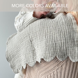 May include: A light gray muslin burp cloth with a scalloped edge and a crinkled texture. The fabric is draped over a shoulder. The text MORE COLORS AVAILABLE is printed in white across the top portion of the image in a semi-transparent bar.