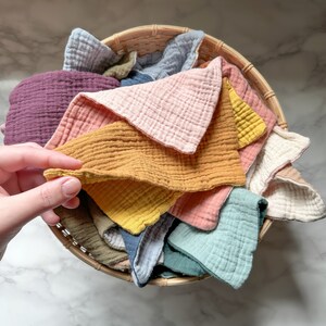 May include: A collection of square muslin cloths in various solid colors including mustard yellow, dusty pink, sage green, plum, light blue, and cream, all neatly arranged inside a round woven wicker basket with a visible textured fabric weave.