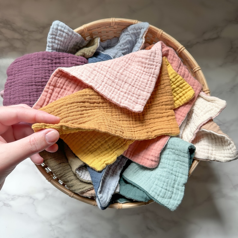 May include: A collection of square muslin cloths in various solid colors including mustard yellow, dusty pink, sage green, plum, light blue, and cream, all neatly arranged inside a round woven wicker basket with a visible textured fabric weave.
