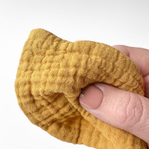 May include: A mustard yellow fabric with a distinct crinkled, multi-layered muslin texture. The material features a grid-like pattern created by the puckered weave of the textile, showing a soft, lightweight, and breathable appearance in a solid color.