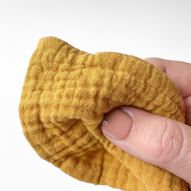 May include: A mustard yellow fabric with a distinct crinkled, multi-layered muslin texture. The material features a grid-like pattern created by the puckered weave of the textile, showing a soft, lightweight, and breathable appearance in a solid color.