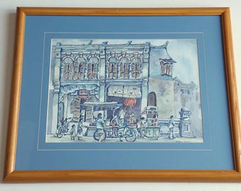May include: Watercolor painting of a street scene in a wooden frame. The artwork depicts a row of buildings with ornate facades, people, bicycles, and a food cart. The color palette is primarily blue and gray, with accents of red.