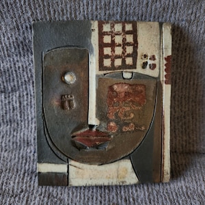 May include: A rectangular ceramic wall plaque featuring a stylised abstract face. The piece uses a palette of charcoal grey, cream, and rust-brown tones. It includes a grid pattern, a single circular eye, and a prominent red-brown lip detail.