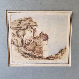 May include: A sepia-toned print depicting a castle on a cliff, with a tree to the left. The castle features multiple towers and a red roof. The print is framed with a black border and set against a grey background. The text "The Castle of Chillon" is visible.