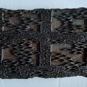 May include: A rectangular dark brown wooden printing block featuring a deeply carved, intricate pattern of repeating leaf-like shapes and textured cross-hatched lines. The surface displays a weathered, aged finish with visible wood grain.