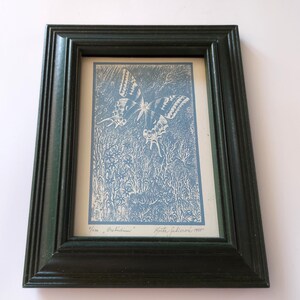 Peut inclure: Un cadre rectangulaire vert foncé contenant une impression bleue et blanche représentant un papillon survolant un feuillage dense. L'impression porte le texte manuscrit "1/100 Delirium" en bas à gauche et une signature avec l'année "1995" en bas à droite.