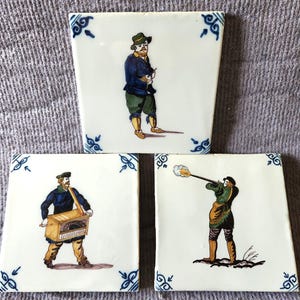 May include: Three square ceramic tiles with a white background and blue corner accents. Each tile features a hand-painted figure: one person standing, one person playing a wooden barrel organ, and one person firing a long rifle with smoke.