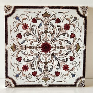 May include: A square ceramic tile with a white background and a brown border. The tile features a symmetrical design of stylised floral motifs in shades of red, brown, and blue.