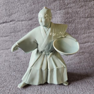 May include: A pale blue-green glazed ceramic figurine depicting a person in a traditional robe with wide sleeves and pleated trousers, captured in a dynamic stance while holding a small, shallow bowl in one hand and extending the other arm.
