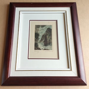 May include: A framed etching featuring a sailboat on water near a rocky cliffside. The artwork is set within a cream-coloured mat and a dark brown wooden frame. The print includes a handwritten title and signature at the bottom of the image area.