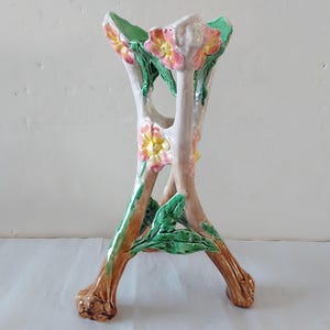 May include: A ceramic plant stand in a tree branch design. The stand is painted with a light pink and brown base, with green leaves and yellow and pink flowers. The stand has a three-legged base and a top opening.