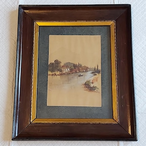 May include: A framed watercolour painting showing a river scene with buildings and trees on the banks. The artwork is set within a dark brown wooden frame with a gold inner border. The painting has a vintage aesthetic.
