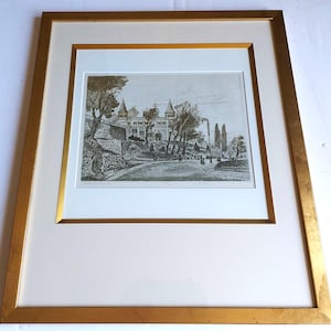 May include: A framed black and white etching depicting a landscape with a large building featuring two towers, surrounded by trees and a winding path. The artwork is encased in a wide, metallic gold frame with a thick, off-white matting border.