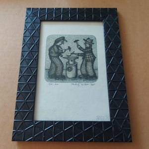 May include: A rectangular black picture frame with a geometric triangular pattern containing a black and white print of two figures working at an anvil. The print includes handwritten text at the bottom, including the date 1985 and a signature.