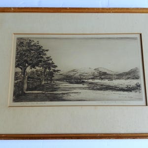 May include: A framed black and white etching depicting a landscape with a tree-lined road, a body of water, and distant mountains. The artwork is enclosed in a light-coloured mat and a simple wooden frame with visible wear on the edges.