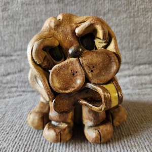 Peut inclure: Une figurine de chien en céramique brune avec une finition texturée et rustique. La sculpture présente de grandes oreilles tombantes, des yeux sombres incrustés, un museau proéminent avec de petites narines pointillées et une longue langue claire.