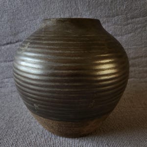 May include: A rounded ceramic vase with a dark brown, metallic-toned glaze and a horizontal ribbed texture covering the upper body. The base of the vase is unglazed, revealing a rough, earthy brown clay texture that contrasts with the smooth finish.