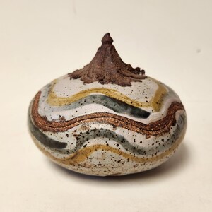 May include: A ceramic jar with a bulbous body and a textured, dark brown top. The jar is decorated with wavy bands of grey, gold, and brown, with speckled details. A unique, handcrafted decorative item.