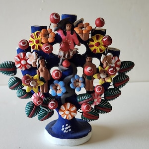 May include: A vibrant ceramic tree sculpture, hand-painted with figures, flowers, and red ball accents. The tree is blue, with a white base, set against a plain background. A unique decorative piece, ideal for display.