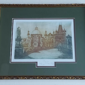 May include: A framed print depicting a European city scene, with a bridge, buildings, and a dome. The artwork uses muted blues, browns, and beige. The frame is gold with decorative detailing, and the print is mounted in green.