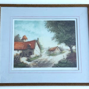 May include: Framed watercolour print of a European village scene. The artwork depicts a stone house with a red tile roof, a dirt track, and a tree. The frame is gold-coloured, and the mount is light blue.