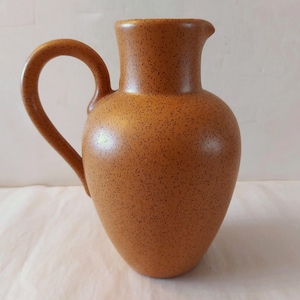 May include: A brown ceramic jug with a handle and a spout. The jug has a speckled texture and a rounded body. The handle is curved and attached to the side. The spout is small and angled.