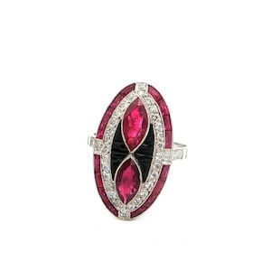 May include: An oval-shaped ring with a silver band. The ring features a central design with two ruby-colored marquise-cut stones, framed by black onyx and surrounded by a border of smaller rubies and clear crystals. The ring is set against a white background.