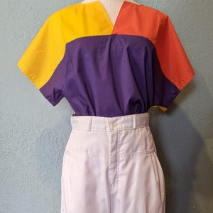 May include: A white pair of high-waisted pants with pleats and a button closure. The pants are paired with a short-sleeved shirt with a yellow, purple, and orange color block design.