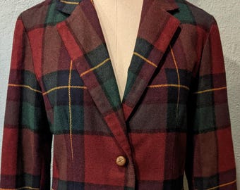 Op de afbeelding: Een vintage geruit pak in tinten rood, groen en marineblauw, tentoongesteld op een mannequin. De jas heeft een sluiting met twee knopen en een reverskraag. De rok is onderaan zichtbaar. Het pak is gemaakt van een geweven stof.