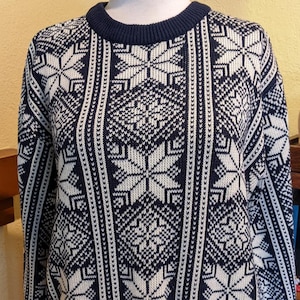 May include: Navy blue and white snowflake patterned sweater. The sweater features a crew neck and ribbed cuffs and hem. The design includes vertical stripes and large snowflake motifs, creating a classic winter look.