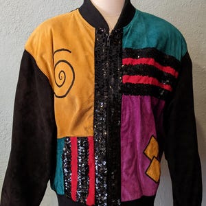 May include: A vintage patchwork jacket in a vibrant colour scheme. The jacket has panels of mustard yellow, teal, purple, and black suede, with black sequin detailing and a spiral design. It is displayed on a mannequin.