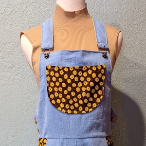 Könnte beinhalten: Hellblaue Jeans-Latzhosen mit einer braunen und gelben Blumendrucktasche. Die Latzhosen haben verstellbare Träger und eine weite Beinweite.