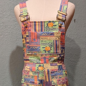 Op de afbeelding: Een brede broek met bretels met een patchwork-ontwerp in verschillende kleuren, waaronder roze, geel, groen, blauw en oranje. De stof heeft een patroon van sinaasappels, bloemen en geometrische vormen. De broek heeft verstelbare schouderbanden met messing gespen.