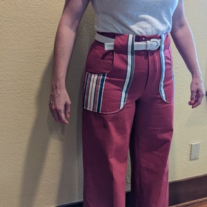 Puede incluir: Un pantalón de pierna ancha de color burdeos oscuro con un cinturón blanco y un detalle de tela a rayas en los bolsillos.