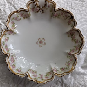 May include: A white porcelain scalloped dish featuring a delicate pink floral pattern with green leaves. The edges are trimmed in gold, and a small gold bow detail is centered at the top. A small floral cluster is positioned in the center of the dish.