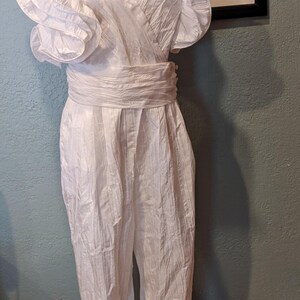 May include: A vintage white jumpsuit with a wrap-style bodice and wide-leg pants. The shoulders feature ruffled detailing, and a matching belt cinches the waist. The garment is displayed on a mannequin, showcasing its design and texture.