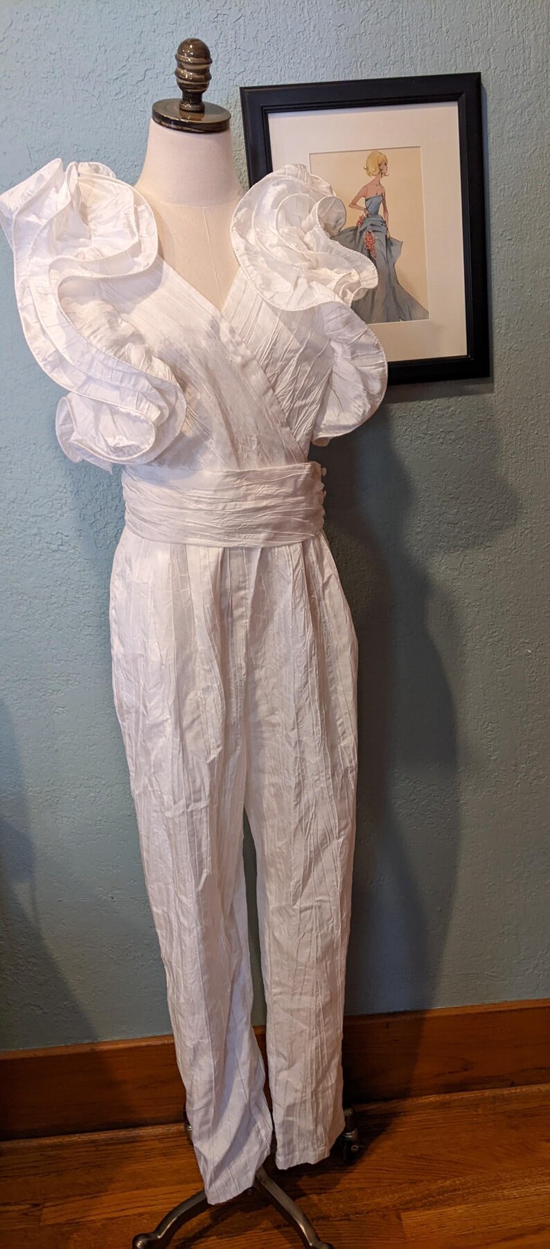May include: A vintage white jumpsuit with a wrap-style bodice and wide-leg pants. The shoulders feature ruffled detailing, and a matching belt cinches the waist. The garment is displayed on a mannequin, showcasing its design and texture.