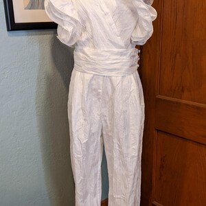 May include: A white jumpsuit with a V-neckline and ruffled sleeves. The waist is cinched with a matching fabric belt. The jumpsuit is displayed on a mannequin. A framed picture hangs in the background.
